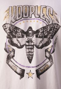 Bílý tričko s potiskem černého můry, s textem "KOOPLES" nad ním, a kruhovým designem s fialovými a zlatými akcenty, včetně černé stuhy.