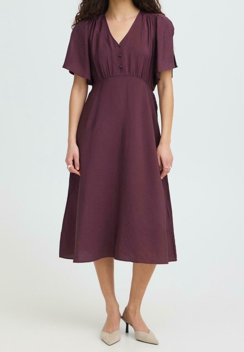Femme portant une robe violet foncé longueur genou avec de courtes manches volantées et des talons beige à bout pointu, debout devant un fond uni.