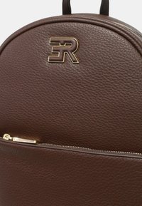 Zaino in pelle marrone con finitura testurizzata, caratterizzato da un logo dorato prominente e una tasca anteriore con zip. Forma arrotondata con spallacci robusti.