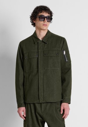 Jonge man draagt een donkergroene jas met knoopsluitingen op de borstzakken en een ritsdetail op de mouw, gecombineerd met bijpassende broek en zwarte zonnebril.