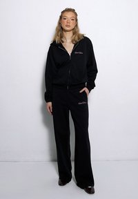 Czarna bluza z zamkiem błyskawicznym z haftowanym logo, w połączeniu z pasującymi czarnymi spodniami szerokimi. Gładka tekstura materiału, minimalistyczny design, luźny krój.