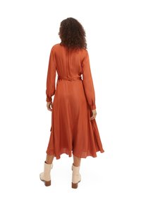 Robe à manches longues, rouge-orange, avec une taille à nouer, jupe fluide et texture lisse. Paire de bottines beige clair aux talons carrés.