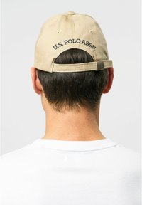 Beige baseball cap with adjustable strap, featuring dark embroidered text "U.S. POLO ASSN." on the back. Worn by a person wearing a white shirt.