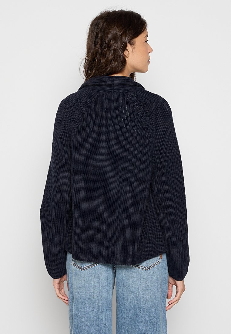 Eine Frau mit welligem braunem Haar trägt einen navy-blauen, grob gestrickten Pullover und hellblaue Jeans, gezeigt von hinten vor einem schlichten Hintergrund.