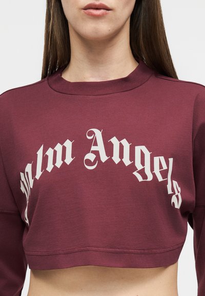 Γυναίκα που φορά ένα κοντό μπουφάν σε μπορντό χρώμα με την επιγραφή "Palm Angels" γραμμένη με λευκά γράμματα σε παλιά αγγλική γραμματοσειρά στο στήθος.