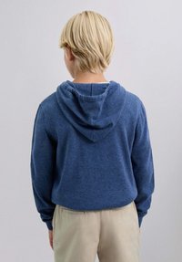 Sudadera azul con capucha, de tejido texturizado y capucha ajustable. La parte trasera presenta un ajuste relajado y se combina con pantalones de color claro.