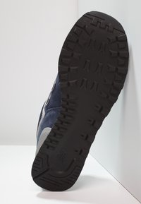 Blå mockaskor med en texturerad svart gummisula, utrustad med ett räfflat mönster och kontrasterande grå detaljer längs sidorna.