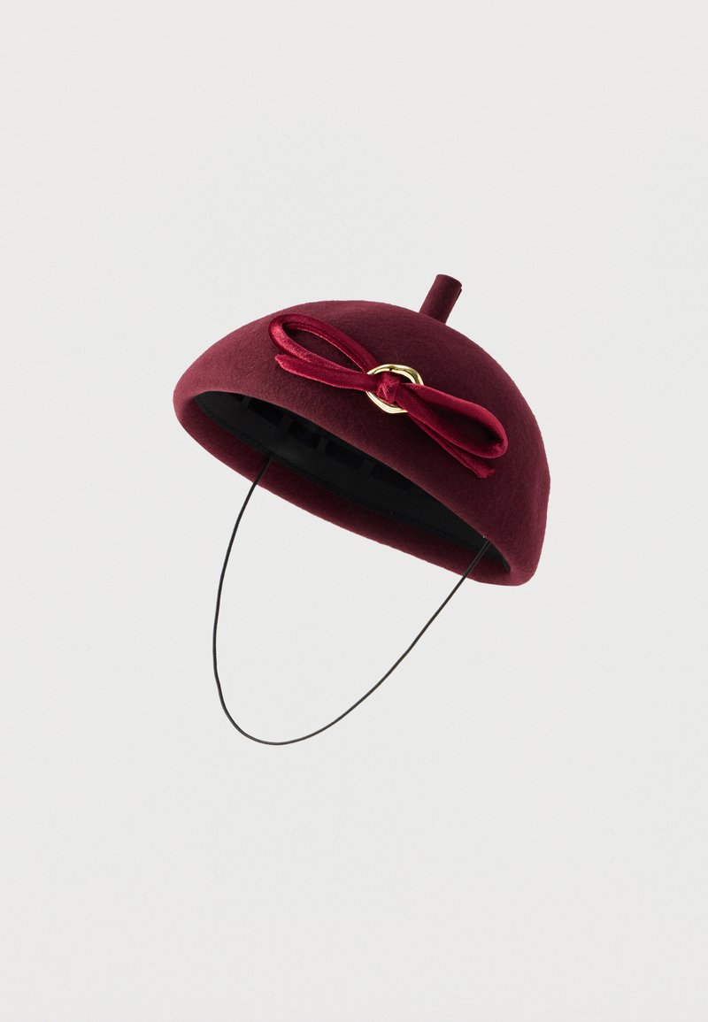 Bordowy filcowy kapelusz o zaokrąglonym kształcie, zdobiony atłasową kokardą i małą pętelką na górze, przymocowany czarną elastyczną opaską.