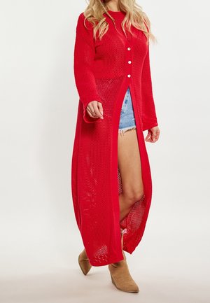 Femme portant un long cardigan rouge en maille ajourée sur un short en jean et des bottines beige, marchant devant un fond uni.