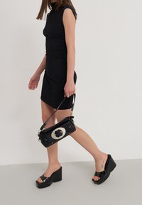 Černá taška přes rameno se stříbrným kruhovým kovovým detailem, doplněná lesklými černými sandály na platformě. Modelka má na sobě přiléhavé černé šaty.