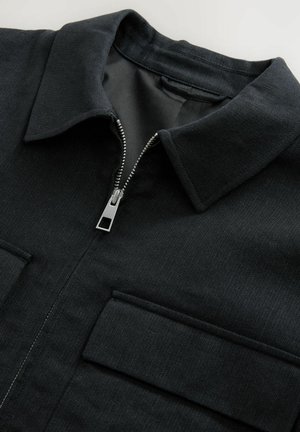 Chaqueta negra con cierre delantero, cuello en punta y dos bolsillos en el pecho; hecha de tela texturizada con detalles de costura visibles.
