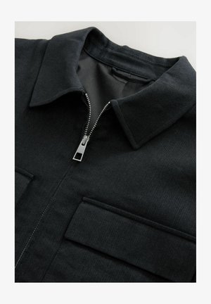 Giacca nera con zip anteriore, colletto appuntito e due tasche sul petto; realizzata in tessuto testurizzato con dettagli di cucitura visibili.