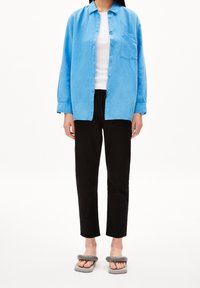 Camicia azzurra con bottoni e maniche lunghe, con taschino sul petto, abbinata a pantaloni neri a gamba stretta e sandali grigi scivolati. Design minimalista, texture liscia.