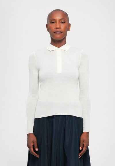 Polo blanc à manches longues côtelé avec un col pointu et une fermeture par boutons, associé à une jupe plissée bleu marine, sur un fond uni.