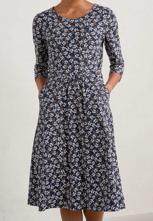 Kobieta ubrana w granatową sukienkę do kolan z białym i brązowym wzorem kwiatowym oraz rękawami do łokcia, z rękami w kieszeniach.
