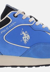 Kék sportcipő texturált bőr és szövet felsőrésszel. Fehér fűzővel, gumitalppal és két póni játékost ábrázoló logóval rendelkezik.