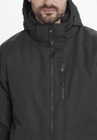 Svart huva jacka i en texturerad tyg som har en framzipper och en bröstficka med dragkedja. Justerbar huva med snodd.