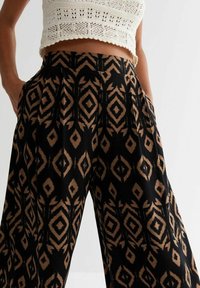 Woman wearing high-waisted black pants with brown geometric diamond pattern and hands in pockets, paired with white crochet crop top.