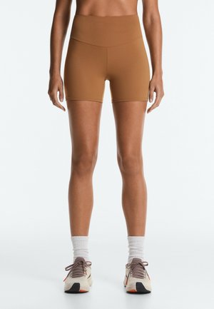 Persoană care poartă pantaloni scurți sport maro cu talie înaltă, șosete albe până la gleznă și adidași albi de alergare cu tălpi negre și portocalii, stând pe un fundal simplu.