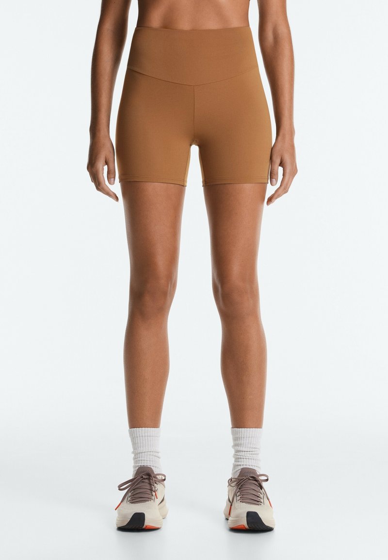 Person trägt braune High-Waist-Sportshorts, weiße Crew-Socken und weiße Laufschuhe mit schwarzen und orangefarbenen Sohlen, steht vor einem einfarbigen Hintergrund.