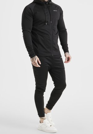 Mandlig figur iført en tætsiddende sort hættetrøje med lynlås og matchende sorte joggers, kombineret med hvide sneakers, stående mod en ensfarvet baggrund.