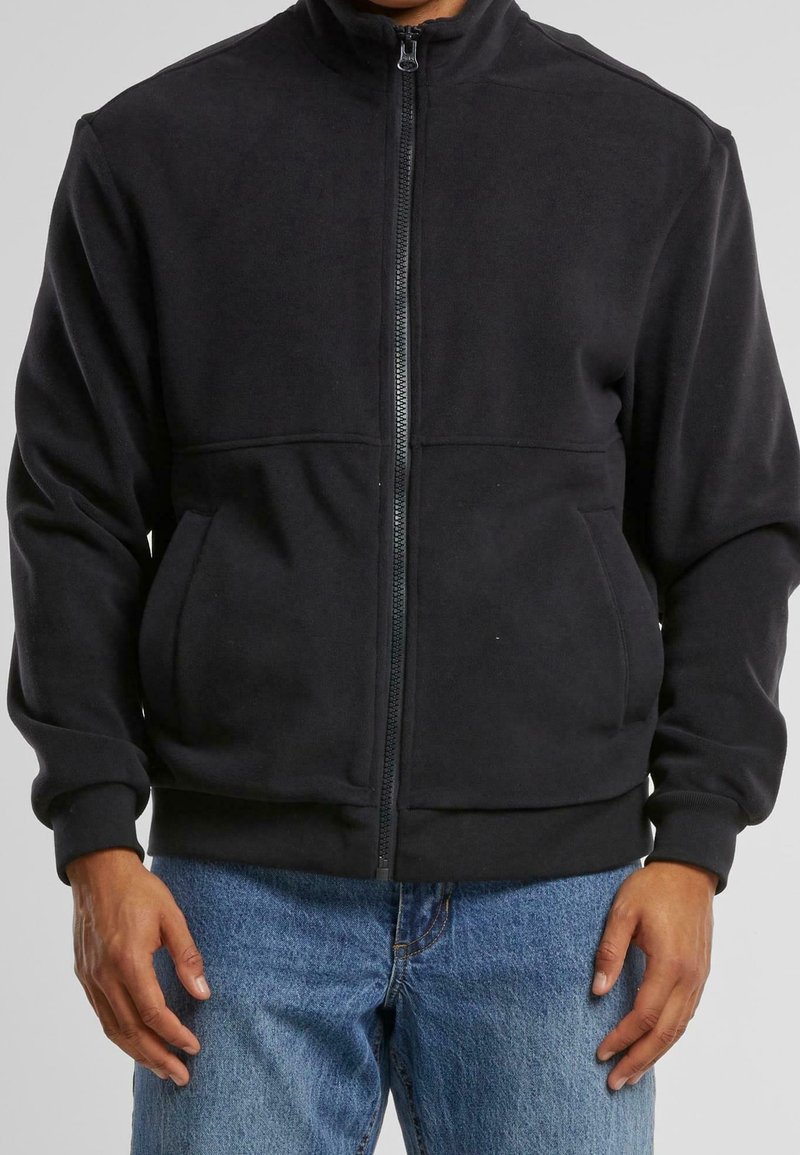 Svart fleecejacka med hög krage, framfickor, ribbade manschetter och en slät yta, i kombination med blå denimjeans.