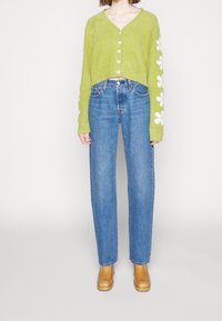 Grön fluffig tröja med V-ringning, prydd med vita blommönster på ärmarna. Kombinerad med blå raka jeans och bruna skor.