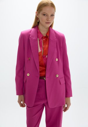 Mujer que lleva un traje cruzado de color rosa brillante con botones dorados y una camisa roja con patrón, de pie frente a un fondo claro y liso.