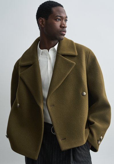 Cappotto doppiopetto in misto lana verde oliva con ampie rever e dettagli sui bottoni, abbinato a una camicia bianca e pantaloni a righe.