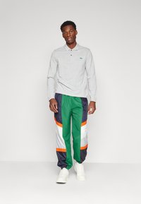 Grijze lange mouw polo met kraag, gecombineerd met groene, marineblauwe en witte trainingsbroeken met oranje accenten en elastische boorden. Witte sneakers.