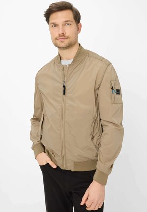 Mann trägt eine beige Bomberjacke mit Reißverschluss, Seitentaschen und Ärmeltaschen, steht mit einer Hand in der Tasche vor weißem Hintergrund.