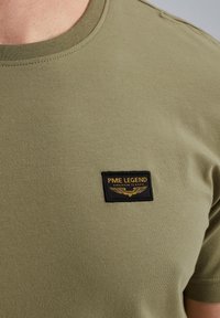 Camiseta de algodón verde oliva con escote redondo y mangas cortas. Presenta una etiqueta bordada en negro que dice "PME LEGEND" con acentos dorados.