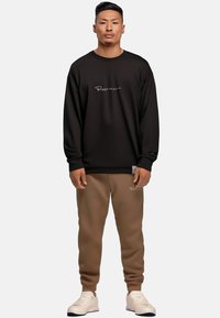 Zwarte sweatshirt met wit logo, gecombineerd met bruine broek met bijpassend logo. Ontwerp is soepel, ontspannen fit, met ribgebreide manchetten.