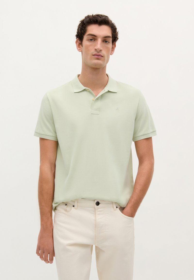 Camisa polo verde claro hecha de un tejido texturizado, con cuello plano, mangas cortas y un pequeño logo en la parte superior izquierda, combinada con pantalones blancos.