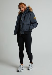 Pelēks polsterēts mētelis ar kažokādas apmali kapucē, ar rāvējslēdzēju un spiedpogu aizdari, divām priekšējām kabatām un logo uzlīmi; kombinēts ar melnajiem legingiem un sudraba sporta apaviem.