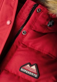 Rote isolierte Jacke mit Reißverschluss und Druckknöpfen, mit einem schwarzen Felsenlogo-Patch. Weicher Kunstfellkragen und strapazierfähige Stoffstruktur.
