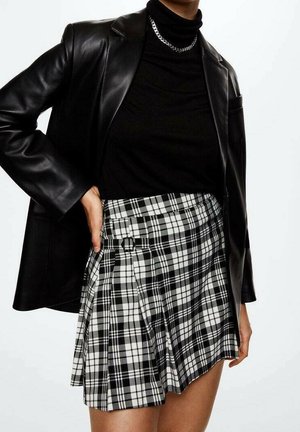 Persona que lleva un blazer de cuero negro, un suéter de cuello alto negro, un collar de cadena plateada y una falda plisada de cuadros en blanco y negro con un detalle de hebilla.