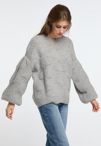 Szürke, oversized pulóver, hullámos mintázattal, hosszú, széles ujjakkal és bordás mandzsettákkal. Kék farmerrel párosítva.