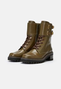 Stivali da combattimento in pelle verde oliva con lacci marroni, fibbie in oro e suola testurizzata. Presentano una cerniera laterale e una punta rotonda.