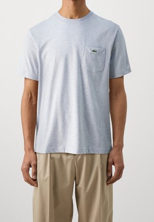 Homme portant un t-shirt gris clair à manches courtes avec une poche poitrine et un petit logo crocodile vert, associé à un pantalon beige.