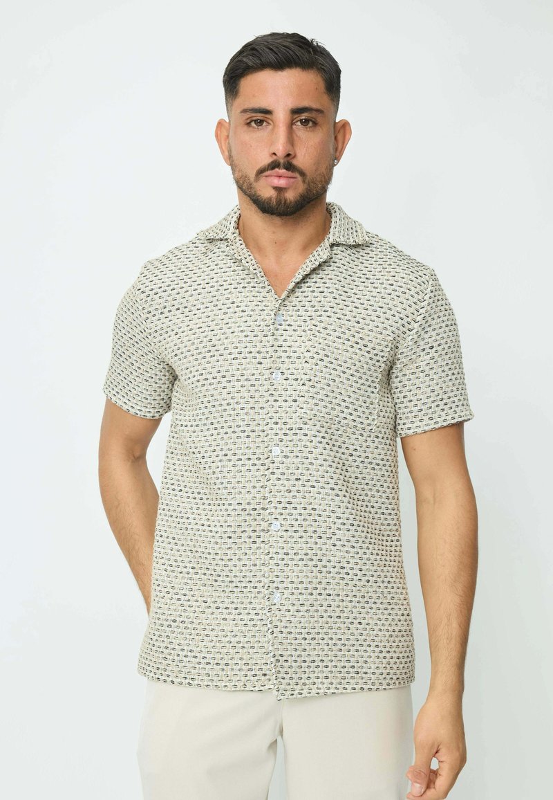 Chemise à manches courtes à boutonnière couleur beige clair, arborant un motif géométrique en bleu marine et vert foncé, avec un col évasé et un tissu lisse.