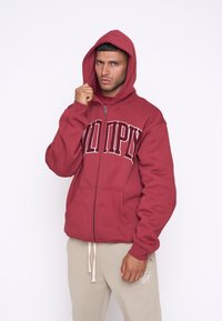 Felpa rossa con zip e grande logo bianco, dotata di cappuccio e tasche frontali. Indossata con pantaloni della tuta beige, che mostrano un look casual.