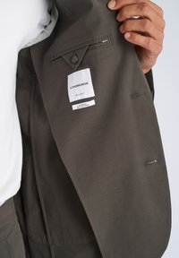 Χέρι που κρατά ανοιχτό σκούρο καφέ σακάκι Lindbergh, δείχνοντας την εσωτερική τσέπη με κουμπί και τις ετικέτες "Κανονική Μεγάλη" και "Superflex Ανακυκλωμένες Ίνες".