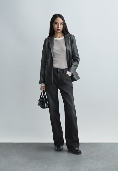 Ženska v črnem usnjenem saku, bež rebrastem topu, črnih širokih hlačah, črnih škornjih, drži majhno črno torbico, stoji na sivi površini.