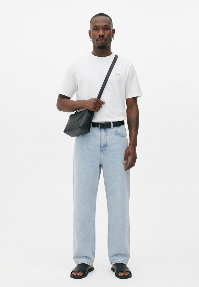 Homme portant un t-shirt blanc, un jean bleu clair, une ceinture noire, un sac bandoulière noir et des sandales noires à enfiler, debout devant un fond blanc.
