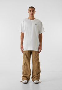Weißes Baumwoll-T-Shirt mit kurzen Ärmeln und einem kleinen schwarzen Logo. Beige Cargo-Hosen mit Taschen und Zugbändern an den Bündchen. Graue Sneakers mit weißen Akzenten.