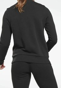 Femme portant un sweat-shirt noir à manches longues et un legging, vue de dos contre un fond uni.