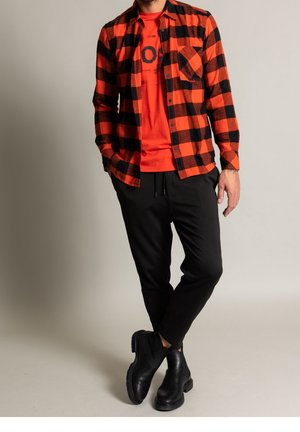 Homme portant une chemise à carreaux rouge et noir par-dessus un t-shirt graphique rouge, pantalon noir et bottines en cuir noir, debout, jambes croisées.