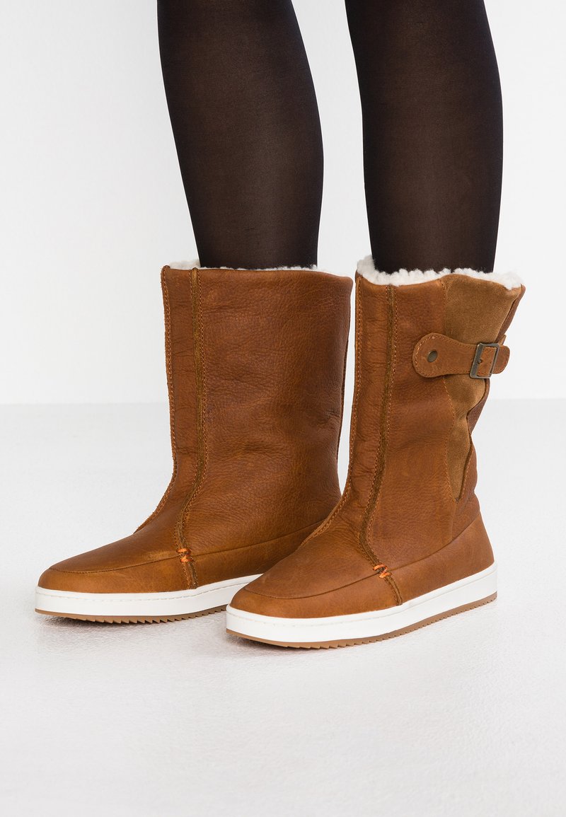 Braune Lederstiefeletten mit einem weichen Fellfutter, einem Schnallenakzent an der Seite und einer weißen Gummisohle. Texturierte Oberfläche mit Nahtdetails.