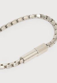 Ασημένιο μεταλλικό βραχιόλι με κρίκους και κυλινδρικό κούμπωμα με σχέδιο, χαραγμένο με την επιγραφή "Calvin Klein" σε λευκό φόντο.